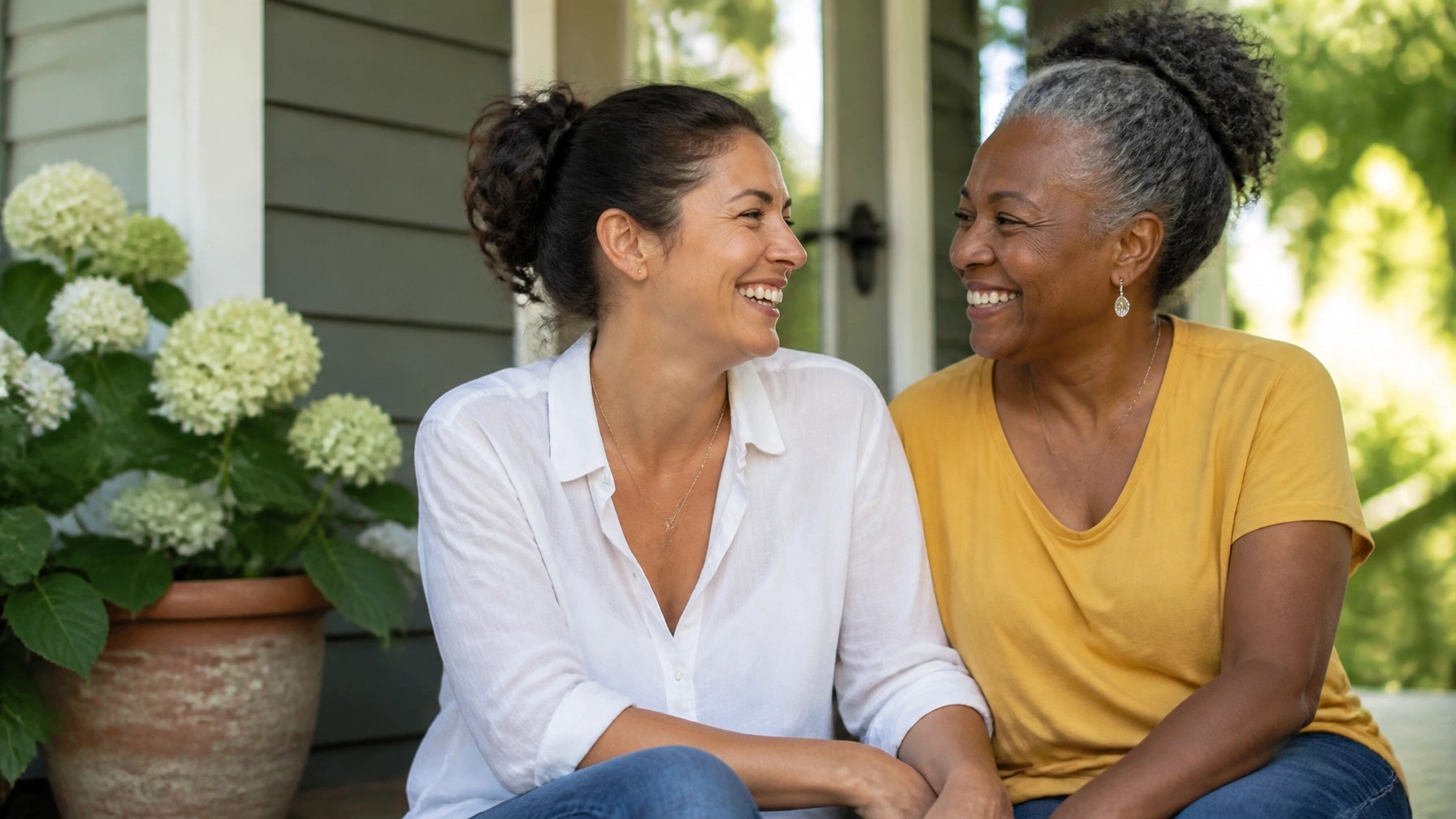 Two diverse women sitting on a sun-drenched porch, sharing a deep conversation and a laugh, looking like lifelong confidants.